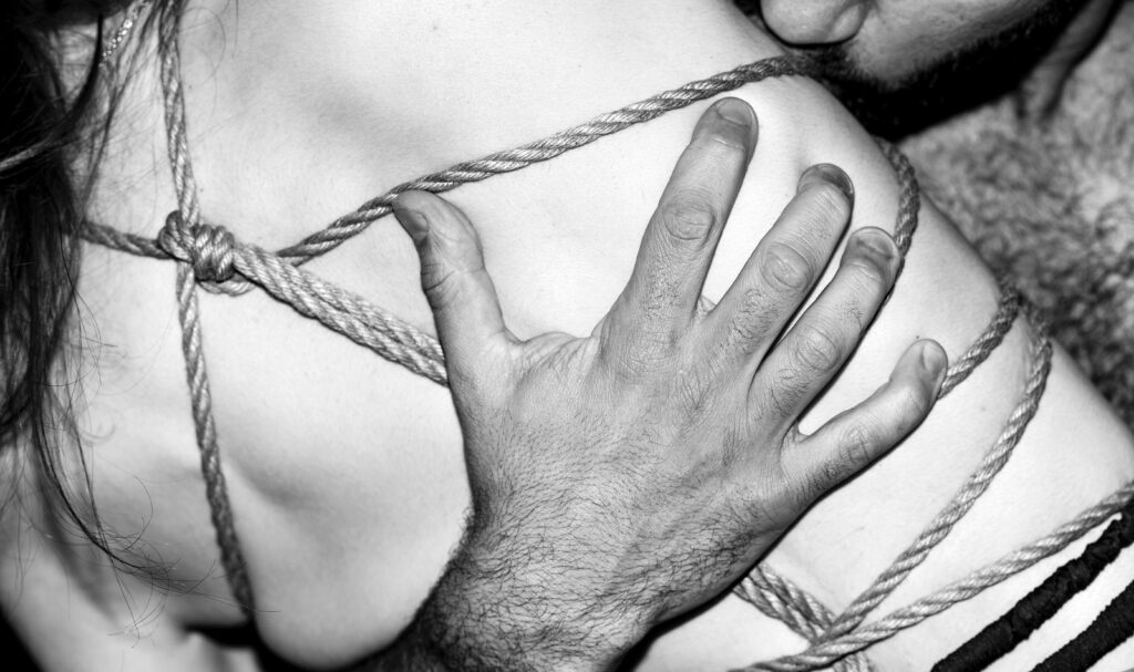 Detaillierte Nahaufnahme einer Bondage-Szene mit Seilen, die kunstvoll um die Schultern einer Person gebunden sind, illustriert durch die präzise Hand eines BDSM Escorts.