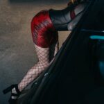 BDSM Escort in verführerischer Pose neben einem dunklen Auto, trägt ein rotes Outfit und Netzstrümpfe, unterstreicht die Kombination aus Eleganz und Provokation in einem urbanen Umfeld
